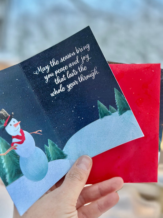 Hand holding a Christmas card with a snowman and festive text against a blurred new england winter storm in boston