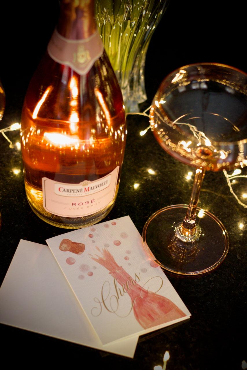 celebration greeting card featuring painted champagne bottle with cork popping and bubbles in the background and elegant script Cheers across the front pictured next to a bottle of pink champagne and pink champagne glass