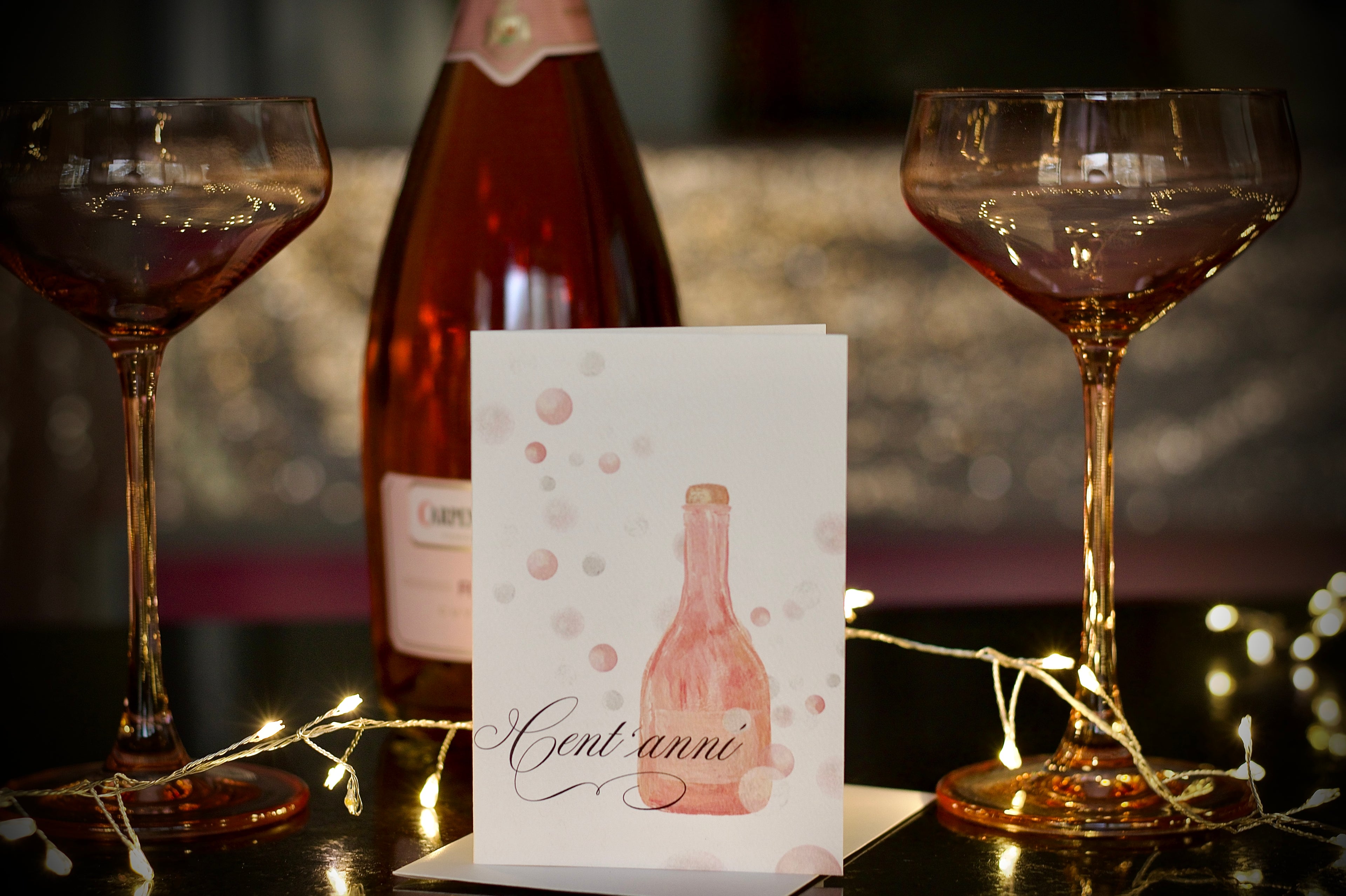 Pink champagne bubbles with pink champagne glasses, featuring a champagne greeting card with Italian greeting cent’anni on the front