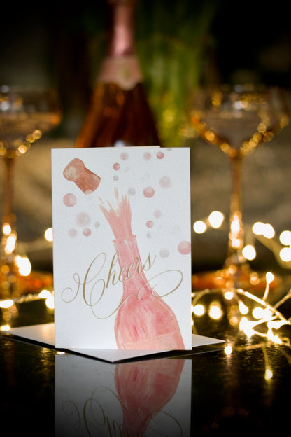 celebration greeting card featuring painted champagne bottle with cork popping and bubbles in the background and elegant script Cheers across the front