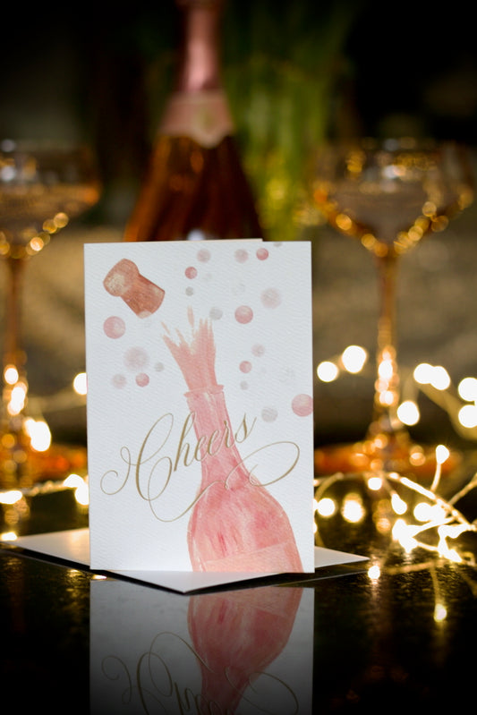 celebration greeting card featuring painted champagne bottle with cork popping and bubbles in the background and elegant script Cheers across the front