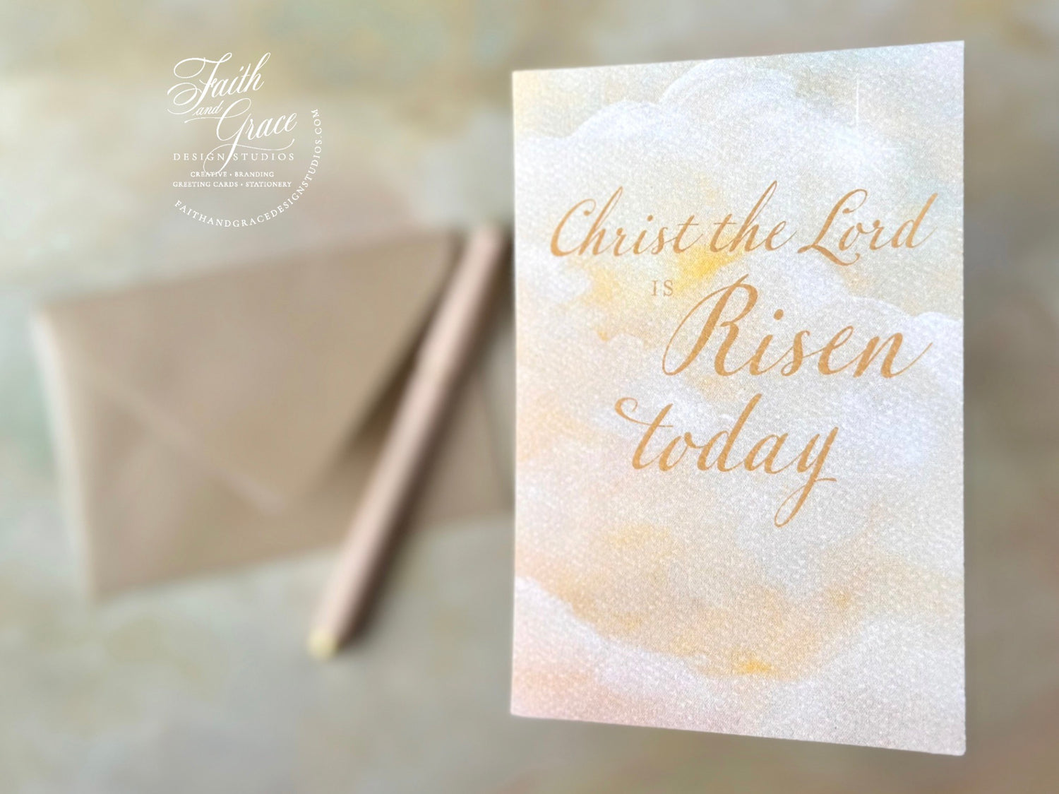 Christ the Lord is Risen Today Easter Card on a warm sunset painted sky with pink pen and sand color envelope blurred in the background.